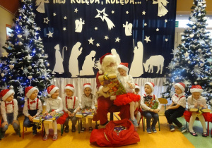 Dziewczynka siedzi na kolanach Mikołaja, ogląda prezent. Wokół siedzi grupa dzieci.